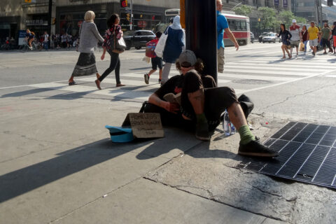 A young homeless man sitting by a pole in a busy street.